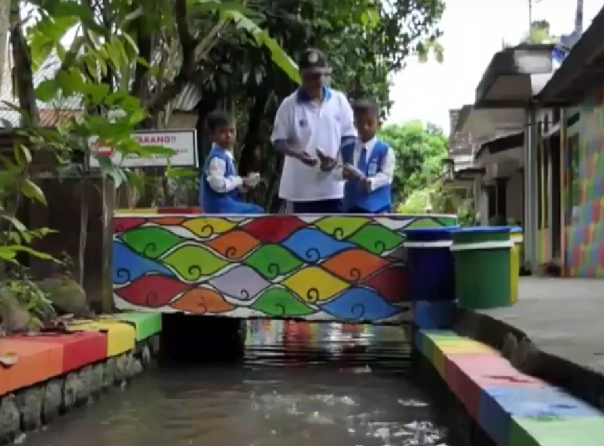 Jembatan warna-warni di atas sungai kecil dengan anak-anak dan pendamping, simbol transformasi lingkungan menjadi ruang belajar komunitas