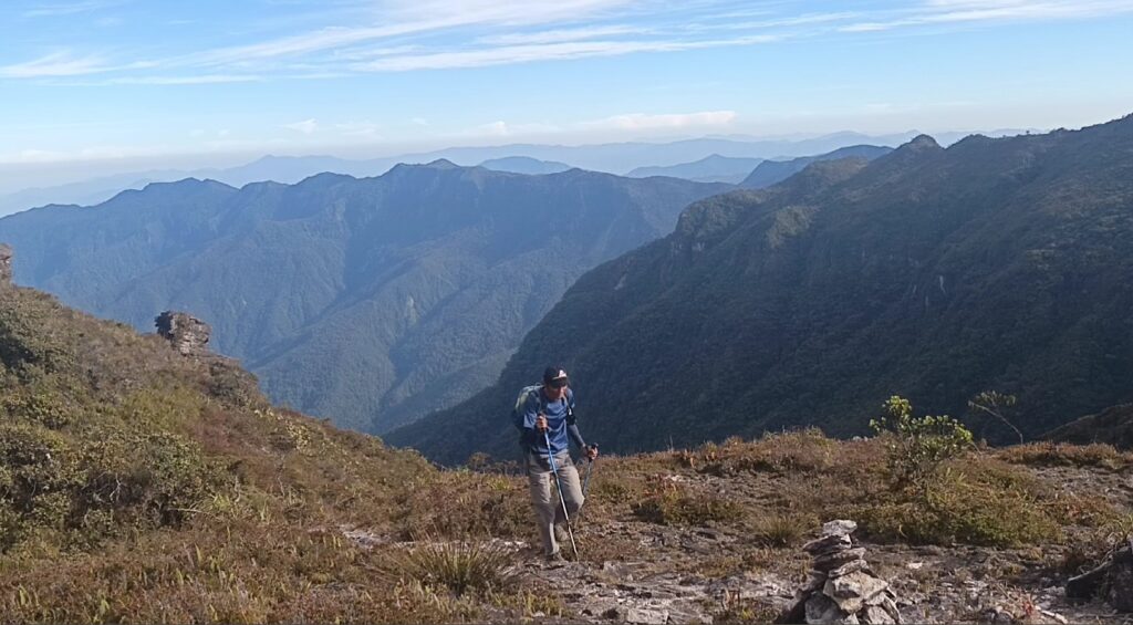Pendaki berjalan di jalur pegunungan Gunung Leuser dengan latar hutan tropis dan langit biru, simbol perjalanan reflektif melawan overthinking